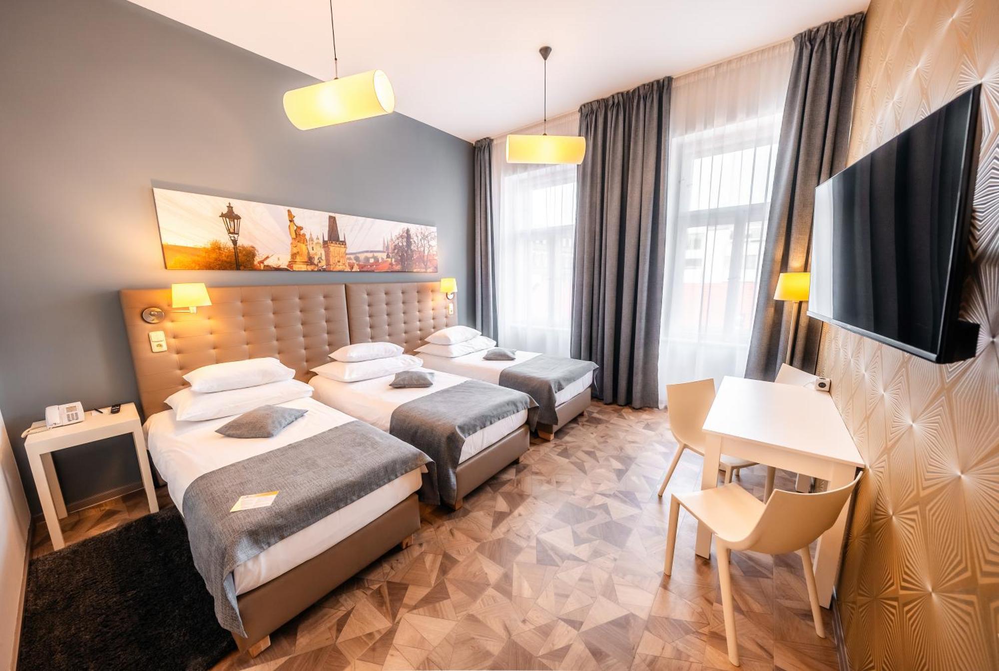 Grandium Hotel Prag Exteriör bild The photo shows a modern hotel room featuring three single beds, each with white bedding and gray throws. The walls are painted gray, and artwork depicting a cityscape is displayed above the headboard. There are two stylish pendant lights hanging fro