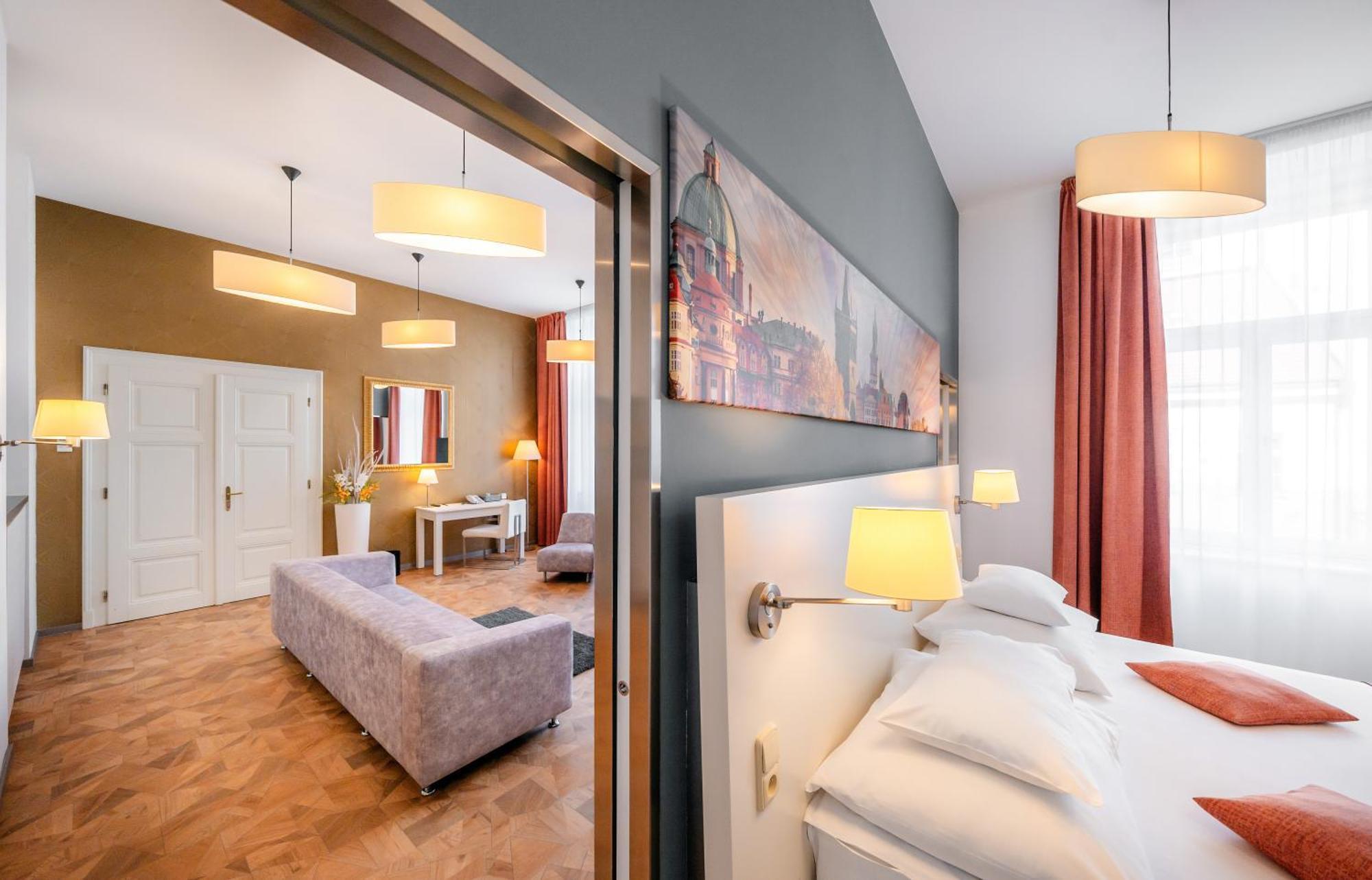 Grandium Hotel Prag Exteriör bild The photo shows a stylish hotel room with modern decor. In the foreground, there is a bed with white pillows, and wall-mounted lamps provide soft lighting. Behind the bed, there's an open doorway leading to a living area featuring a light-colored sof